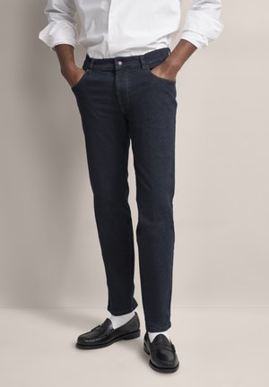 Dunkelblaue Jeans mit schmaler Passform, ausgestattet mit einem regulären Bund und Taschen. Tragen mit schwarzen Loafers und weißen Socken.