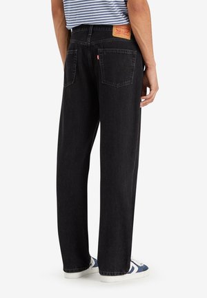Personne portant un jean noir droit avec poches arrière et une étiquette de marque en cuir marron, assortie à des baskets bleues et blanches.