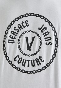 Chemise grise arborant un logo noir circulaire avec le texte "VERSACE JEANS COUTURE" et un "V" stylisé central, entouré de motifs de chaînes.