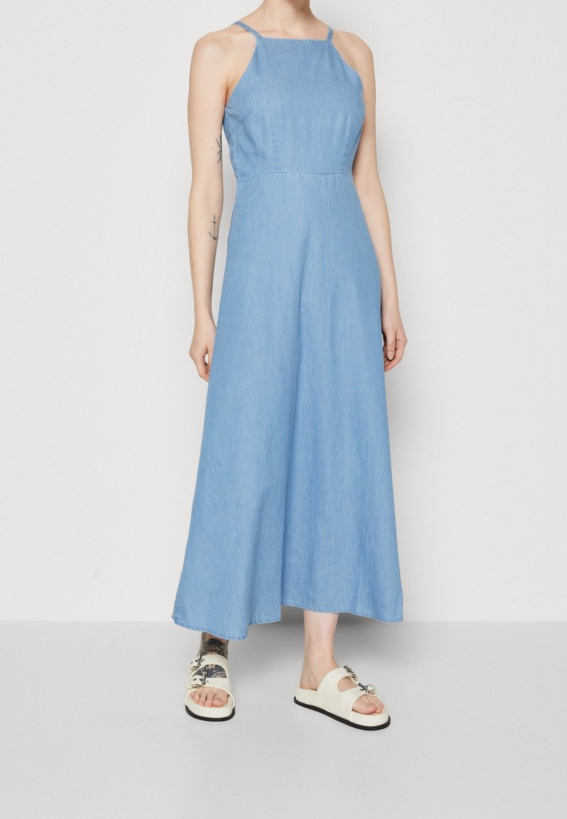 Vaaleansininen farkkukangas maxi-mekko, jossa on ohuet olkaimet, vartalonmyötäinen yläosa ja levenevät helmat. Yhdistetty valkoisiin sandaaleihin, joissa on koristeneula.