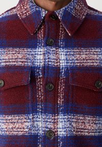 Gros plan d'une veste-chemise en laine à carreaux boutonnée avec des couleurs bleu, bordeaux et blanc et deux poches poitrine avec des boutons noirs.