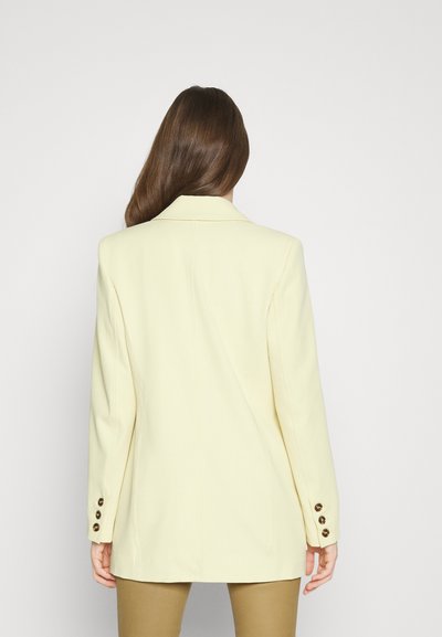 Γυναίκα με μακριά καστανά μαλλιά, φορώντας ένα ελαφρύ κίτρινο σακάκι με κουμπωμένα μανίκια και μπεζ παντελόνι, στραμμένη με την πλάτη στη κάμερα.