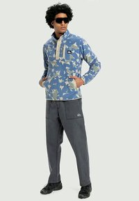 Man wearing blue and beige floral fleece pullover, dark gray pants, black sneakers, and large black sunglasses, standing with hands in pockets.