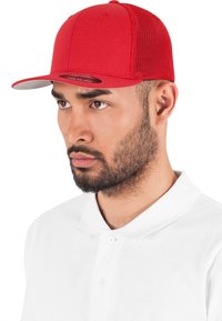 Punane pesapalli müts, millel on tahke etteosa ja võrgu tagaosa, lame visiit ning peene logo allosas, kannab mudel valges polo särgis.