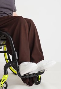 Αναπηρικό αμαξίδιο με μαύρο πλαίσιο και έντονες πράσινες λεπτομέρειες, που διαθέτει ένα άτομο ντυμένο με φαρδιά καφέ παντελόνια και λευκά αθλητικά παπούτσια να ξεκουράζεται στην υποπόδιο.