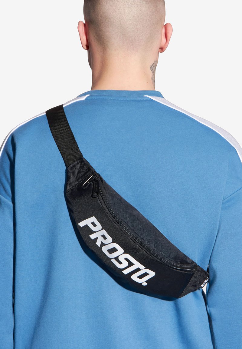 Czarna nerka z napisem "PROSTO" w kolorze białym, noszona na niebieskiej bluzie. Posiada komorę na zamek oraz regulowany pasek.