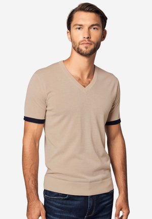 Mann trägt beige kurzärmeligen V-Ausschnitt-Pullover mit schwarzem Besatz an den Bündchen und dunkelblaue Jeans, steht vor weißem Hintergrund.