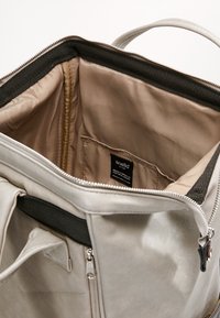 Sac avec doublure intérieure beige, bordure noire, une poche intérieure avec une étiquette de marque visible, et une fermeture éclair argentée ouverte sur le dessus.