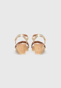 Sandales en liège beige à talon compensé avec des brides métalliques dorées autour de la cheville et des accents décoratifs noirs. Talon en liège texturé, design à dos ouvert.