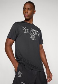 Crna majica kratkih rukava s bijelim "Yankees" logotipom i NY emblemom. Glatki materijal s opuštenim krojem, u kombinaciji s odgovarajućim crnim kratkim hlačama.