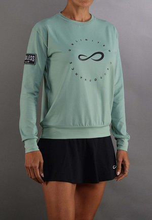 Donna che indossa una felpa verde chiaro con testo "Unlimited Performance" e gonna nera, in piedi davanti a uno sfondo grigio uniforme.