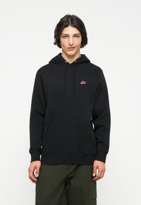 Juodas hoodi su priekinėmis kišenėmis, su mažuoju rožiniu Nike logotipu kairėje krūtinės pusėje. Reguliuojama kapučino su raišteliais ir gumbinės rankovės.