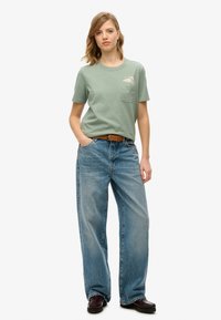 Grøn bomulds t-shirt med brystlomme og grafisk design, parret med løstsiddende lyseblå denimjeans og brune sko.