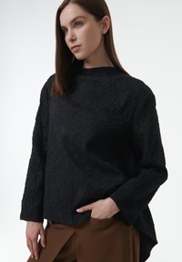 Top negro de manga larga con textura, que presenta un cuello alto y un ajuste relajado, combinado con pantalones marrones. Detalle sutil de arrugas en toda la prenda.