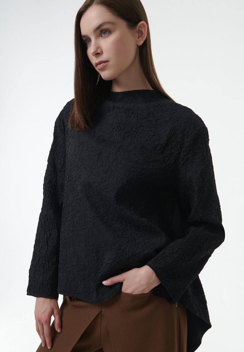 Top negro de manga larga con textura, que presenta un cuello alto y un ajuste relajado, combinado con pantalones marrones. Detalle sutil de arrugas en toda la prenda.