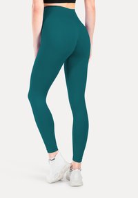 Leggings de talle alto en color verde azulado, fabricados con un tejido elástico, que presentan una textura suave y un diseño ajustado, combinados con zapatillas deportivas blancas.