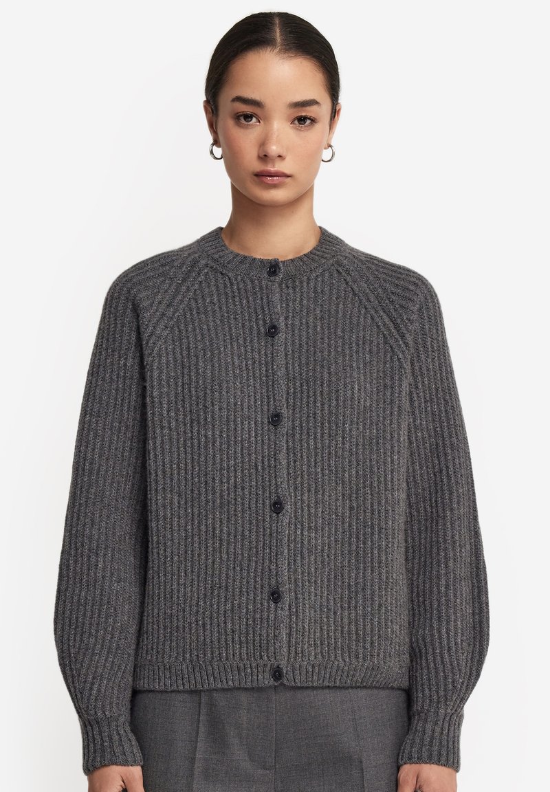 Femme portant un cardigan gris côtelé boutonné à manches longues et col rond, associé à un pantalon gris, debout devant un fond blanc.