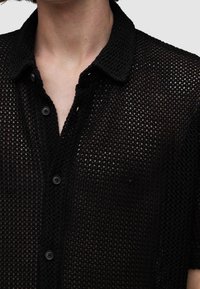 Maglietta nera in rete con collo a camicia, maniche corte e dettagli con bottoni visibili, caratterizzata da un design testurizzato e a maglia aperta.