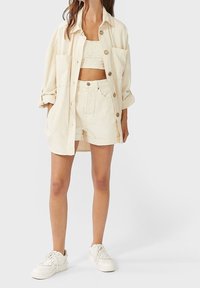 Beżowa, oversize koszula zapinana na guziki, biała koronkowa bluzka typu crop top i pasujące szorty, w zestawieniu z białymi trampkami, wszystko wykonane z miękkiej bawełny.