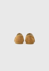 Zapatillas de ante beige tipo slip-on con suela de goma plana, que presentan detalles de costura y un logotipo grabado en el talón.