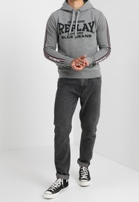 Felpa grigia con cappuccio e grande logo nero "REPLAY", maniche a strisce rosse e nere, abbinata a jeans scuri e sneakers nere.