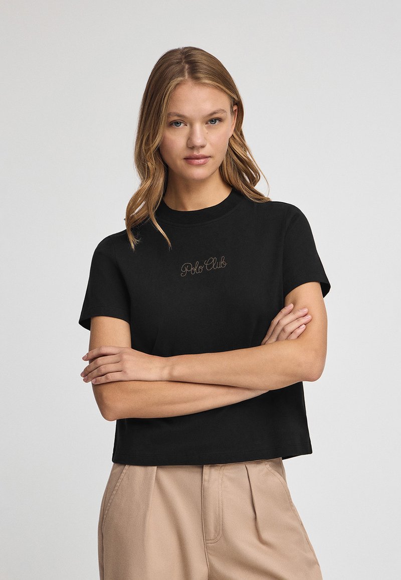 Camiseta negra corta con mangas cortas, que presenta un logotipo bordado en tono "Polo Club" en el pecho. Tejido de algodón suave.