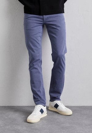 Persona con vaqueros ajustados azules y zapatillas blancas con rayas negras, de pie contra una pared lisa sobre un suelo gris.