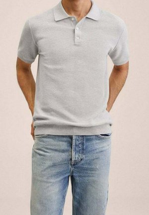 Mand iført lysegrå kortærmet polo, utåget over lyseblå denimjeans med hænderne bag ryggen.