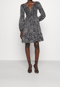 Šaty s černobílým vzorem, dlouhými rukávy, výstřihem do V, staženým pasem a volánovým lemem, vyrobené z lehkého materiálu. Černé sandály na podpatku.