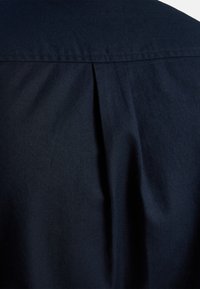 Marineblauwe shirtstof die een achteraanzicht toont met een subtiele plooidetail. De textuur lijkt glad en lichtgeweven, met een gestructureerde kraag.