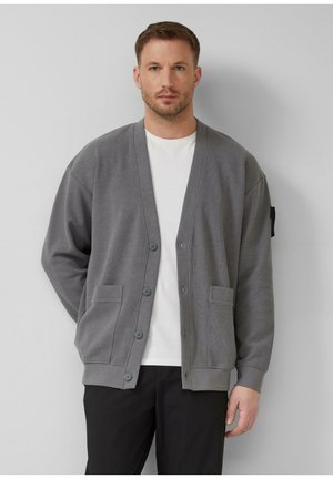 Mann trägt grauen, geknöpften Cardigan über weißem T-Shirt und schwarzer Hose, steht vor hellgrauem Hintergrund, Hände in den Taschen.