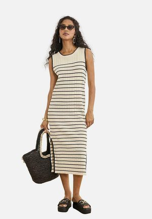 Mujer con vestido midi de punto sin mangas beige con rayas negras, sandalias negras de plataforma, gafas de sol y llevando un gran bolso negro tejido.