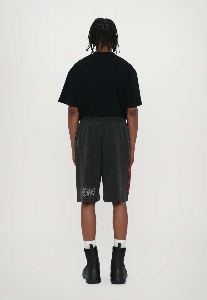 Camiseta negra de gran tamaño, pantalones cortos grises con un gráfico rojo lateral y un logo blanco, combinados con botas negras hasta los tobillos y calcetines blancos. Diseño minimalista.