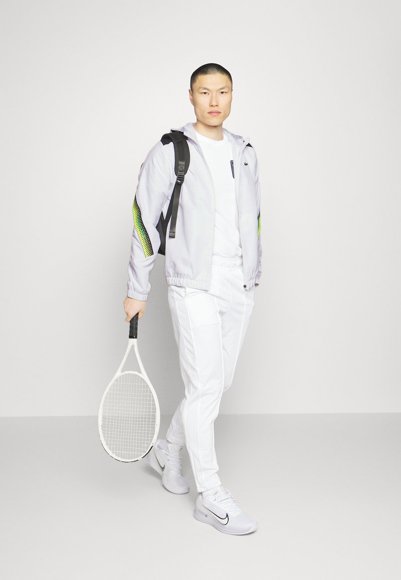 Tenue de tennis comprenant une veste gris clair avec des accents noirs et des détails en maille verte, une chemise blanche, un pantalon et des chaussures blanches. Tenant une raquette de tennis.