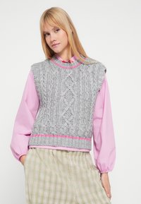 Szary dzianinowy bezrękawnik z wzorem warkocza, z okrągłą różową lamówką, noszony na jasnoróżowej bluzce. Charakteryzuje się luźnym krojem i prążkowanym dołem.