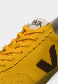Κίτρινο δερμάτινο αθλητικό παπούτσι με καφέ λογότυπο σε σχήμα V, με κίτρινα κορδόνια και υφασμένο πάτο. Διακριτικές ραφές και λεπτομέρειες λογοτύπου στη γλώσσα του παπουτσιού.
