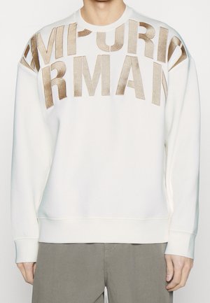 Sweat-shirt blanc à manches longues avec de grandes lettres dorées métalliques formant "ARMANI" sur la poitrine, porté par une personne portant un pantalon beige.