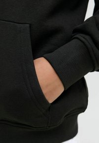 Crna dukserica s kengurusskim džepom, rebrastim manžetama i mekim teksturom. Tkanina izgleda glatko s minimalnim detaljima šavova.