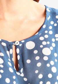 Blusa in tessuto blu con pois bianchi, scollo a chiave con bottone e una texture liscia. Design casual con vestibilità rilassata.