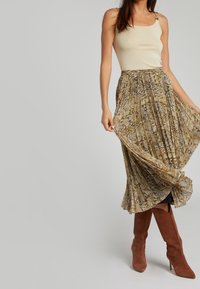 Crème mouwloze top en een bruine, gestreepte maxi rok met plooien. De rok heeft een slangenprint en is gecombineerd met hoge, bruine laarzen.