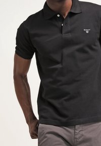 Homme portant un polo noir GANT avec une patte de boutonnage à deux boutons et un pantalon gris, debout devant un fond clair uni.