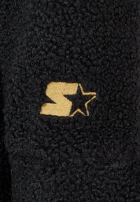 Materialul de fâșie neagră are o suprafață texturată. Un logo auriu cu un "S" și un detaliu în formă de stea este expus proeminent pe material.