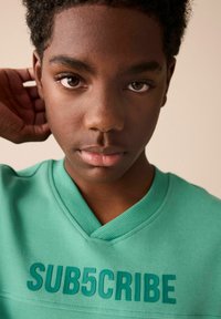 Grøn sweater med v-udskæring, prydet med hævet tekst "SUB5CRIBE" i en mørkere grøn. Nærbillede af ansigt med kort, struktureret hår.