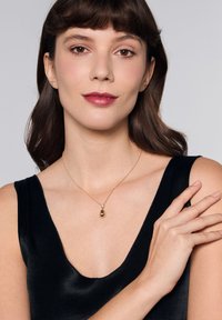 Złoty naszyjnik z zawieszką w kształcie kropli łzy i małym akcentującym kamieniem, noszony przez modelkę w czarnym satynowym topie. Gładka tekstura, delikatny łańcuszek.