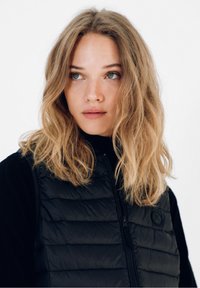 Sort puffer vest med vandret quiltning, der har en høj krave og frontlynlås, lagd over en sort langærmet top.