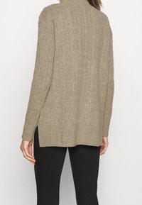 Pull en maille beige avec un motif en torsades, col montant et fentes sur les côtés, fabriqué dans un matériau doux, associé à un legging noir.