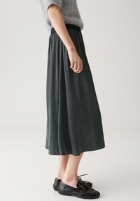Gonna midi di un verde scuro, fluente, con una vita elastica nera; modello abbinato a scarpe nere con lacci. Tessuto morbido e liscio.