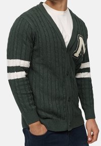 Cardigan a maglia verde scuro con motivo a trecce testurizzato, scollo a V, bottoni frontali e strisce bianche sulle maniche. Presenta un grande accento a lettera color crema.