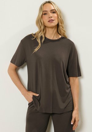 Blonde Frau trägt ein dunkelbraunes, lockeres Kurzarmshirt und passende Hosen, steht mit einer Hand in der Tasche vor einer weißen Wand.