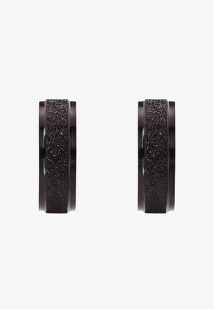 Juodas žiedas su tekstūruota paviršiumi; lygūs kraštai kontrastuoja su šiurkščia, blizgia vidurinė dalimi. Apvalios formos, pagamintas iš metalo ar panašios medžiagos.